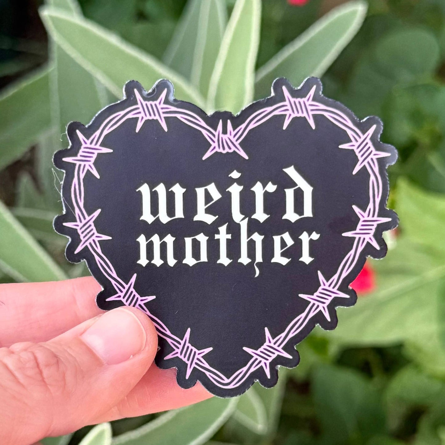 A hand holds a heart-shaped black sticker with a barbed wire border and the text 'weird mother' in white gothic font, set against a background of green leaves and pink flowers.