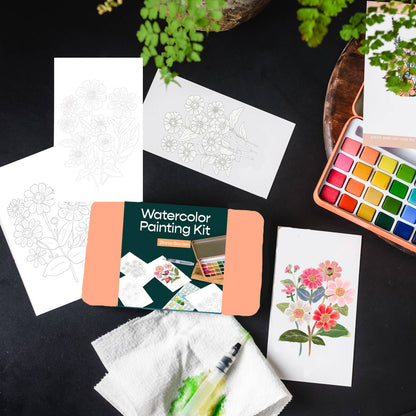 Watercolor painting kit with watercolor paper, paint set, and a plant on a dark surface.