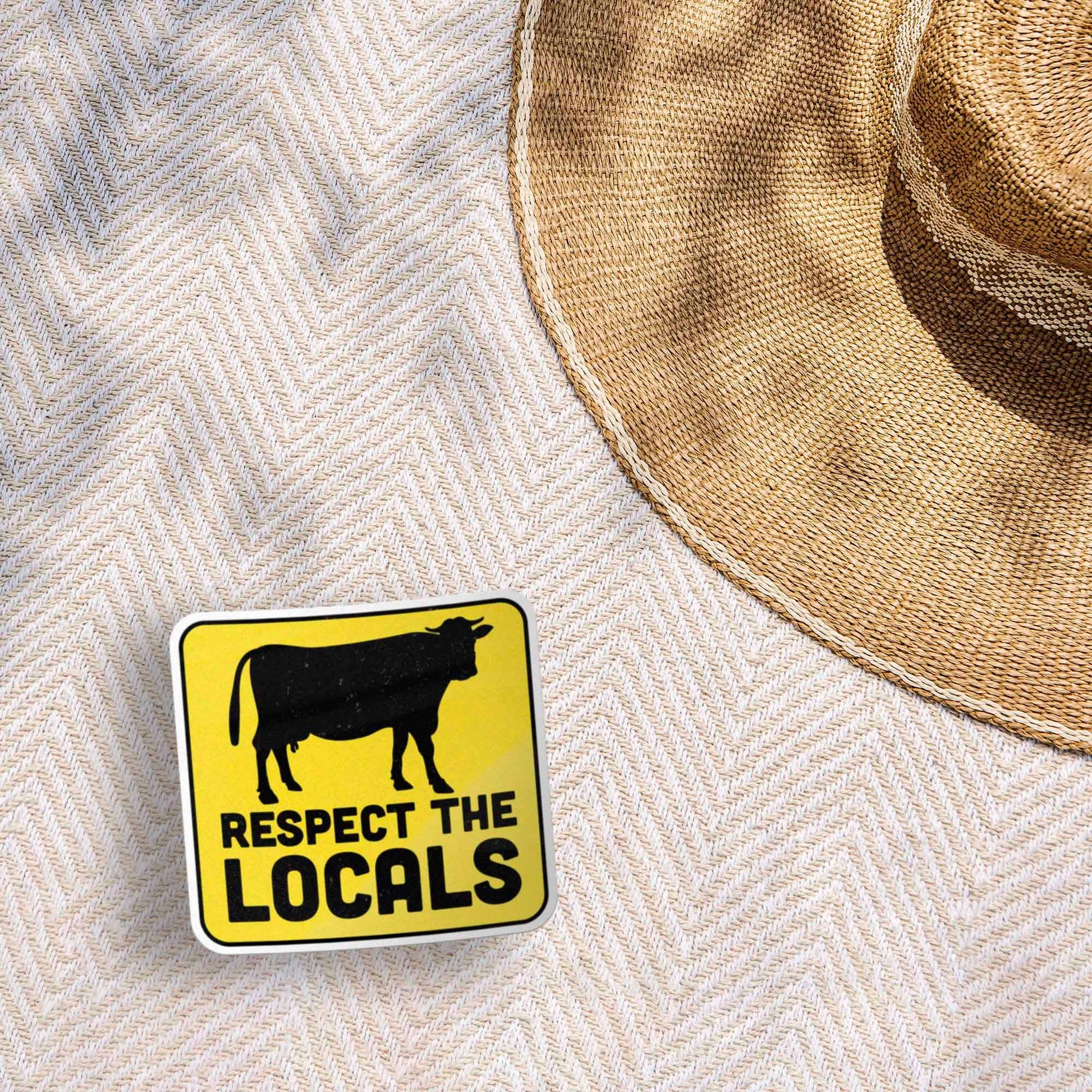 Yellow sign with a cow illustration and 'Respect the Locals' text on a textured surface with a straw hat.