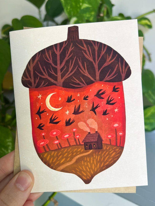 A hand holds a square art print featuring an acorn illustration with a forest canopy on top, a red sky with a crescent moon and flying birds inside, and a small house over a field at the bottom. The print is shown against a blurred background of green leaves.