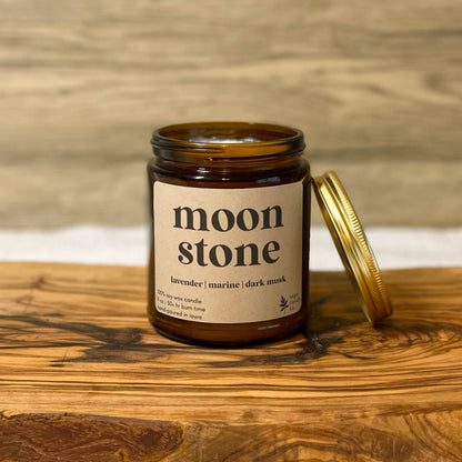 An amber glass jar candle labeled 'moon stone' with its gold metal lid resting beside it on a rustic wooden board, set against a light wood plank background.