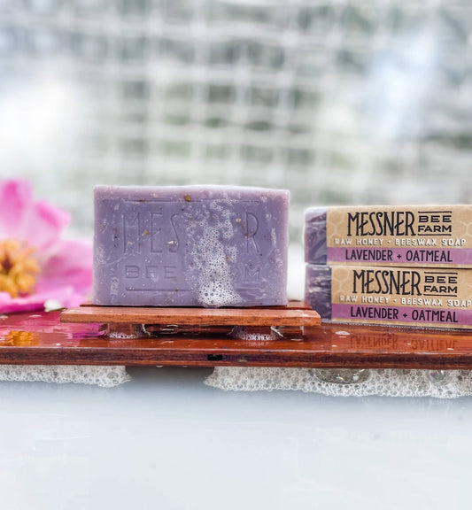 Two bars of Messner Bee Farm lavender and oatmeal soap on a wooden block with a blurred natural background.