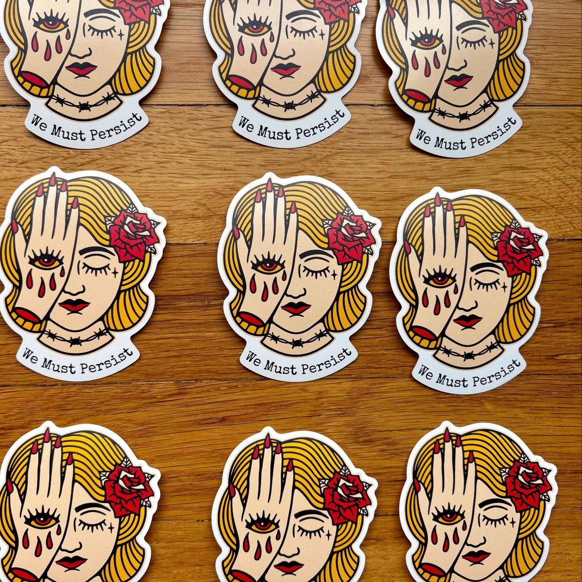 Nine identical die-cut stickers arranged in a 3x3 grid on a wooden surface. The sticker design shows a stylized woman with blonde hair, a red rose, and crossed hands with the text 'We Must Persist' below.