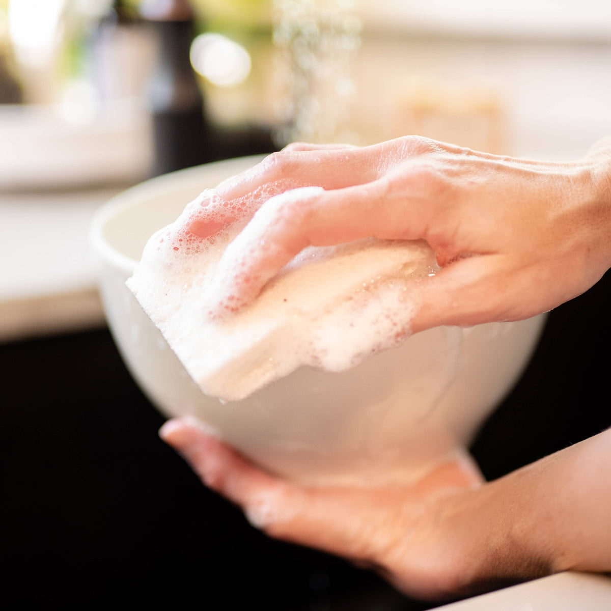 A close-up lifestyle shot of a person's hands washing a white ceramic bowl with a soapy sponge in a kitchen sink.