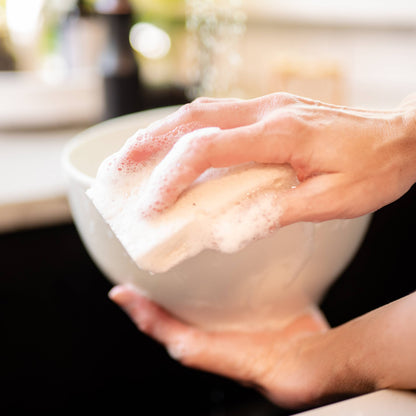 A close-up lifestyle shot of a person's hands washing a white ceramic bowl with a soapy sponge in a kitchen sink.