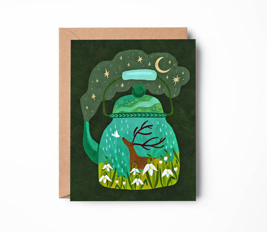 A square greeting card with a kraft envelope, featuring a whimsical illustration of a green teapot containing a deer, snowdrops, and a starry night sky.