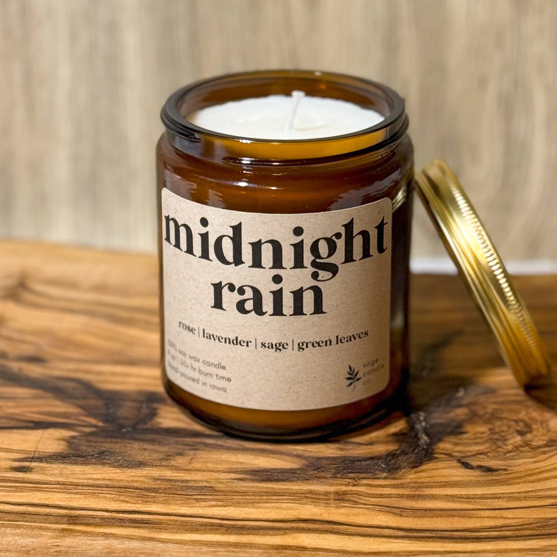 An amber glass jar candle labeled 'midnight rain' with a cream label and a gold lid resting beside it, set on a rustic wooden board against a wood-paneled background.