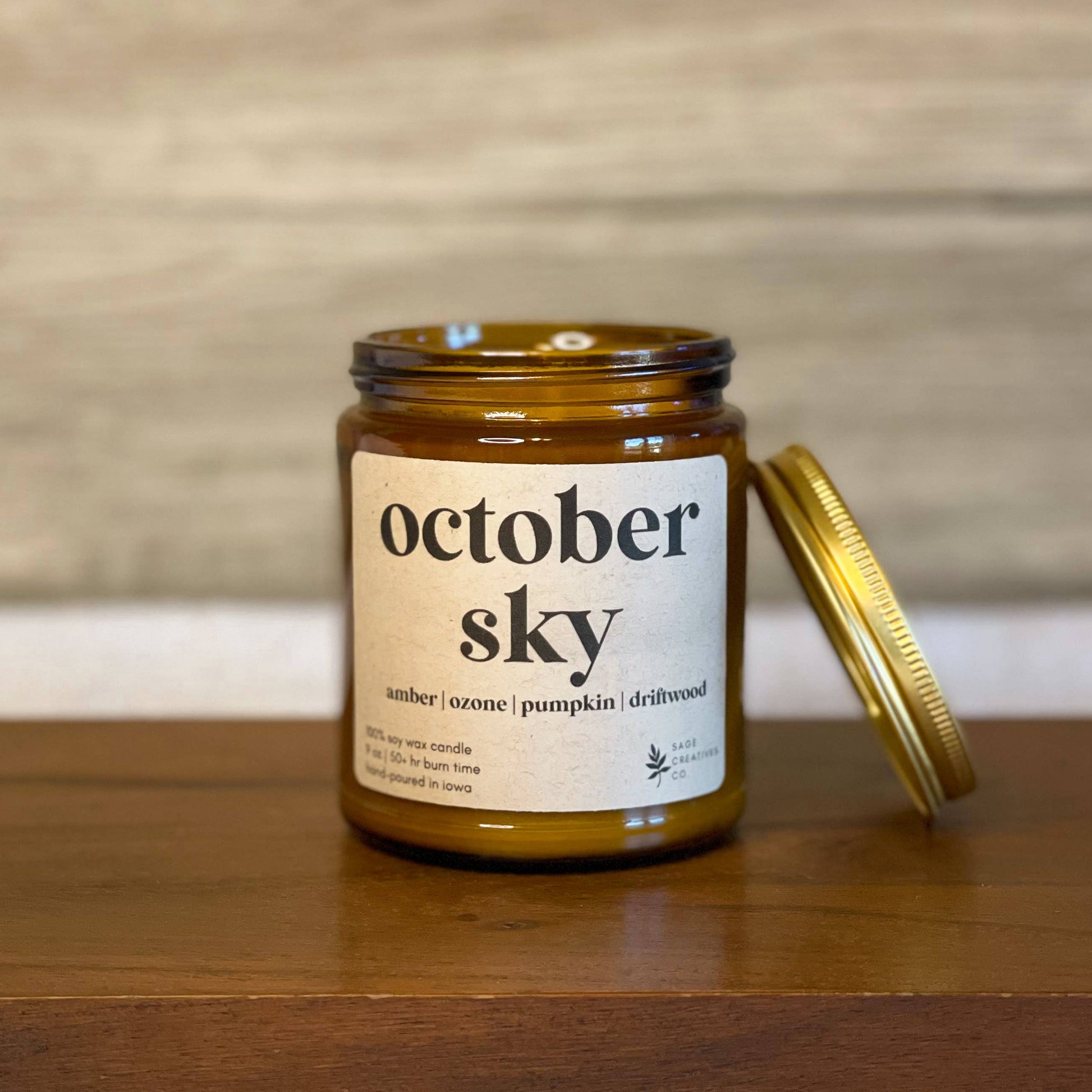 An amber glass jar candle with a cream label reading 'october sky' sits on a wooden surface with its gold metal lid leaning beside it. The background is a softly blurred light wood wall.