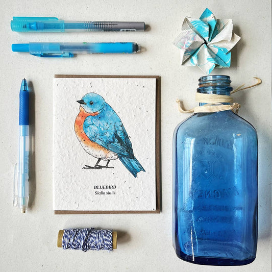 Bluebird illustration card with blue pen, bottle, and decorative items on a light surface