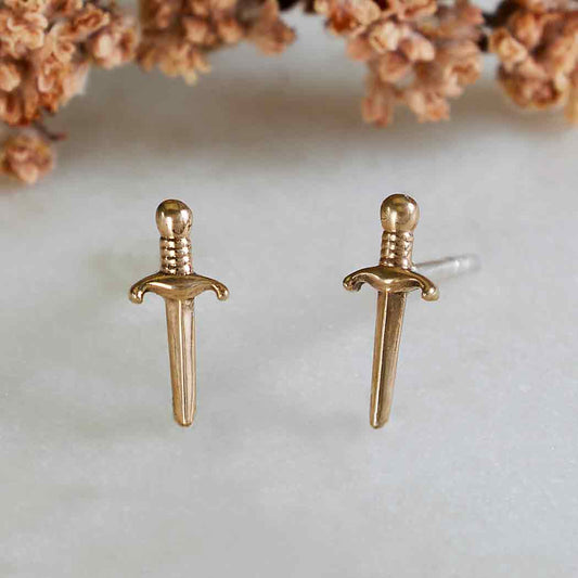 Gold sword-shaped earrings on a light background with dried flowers.