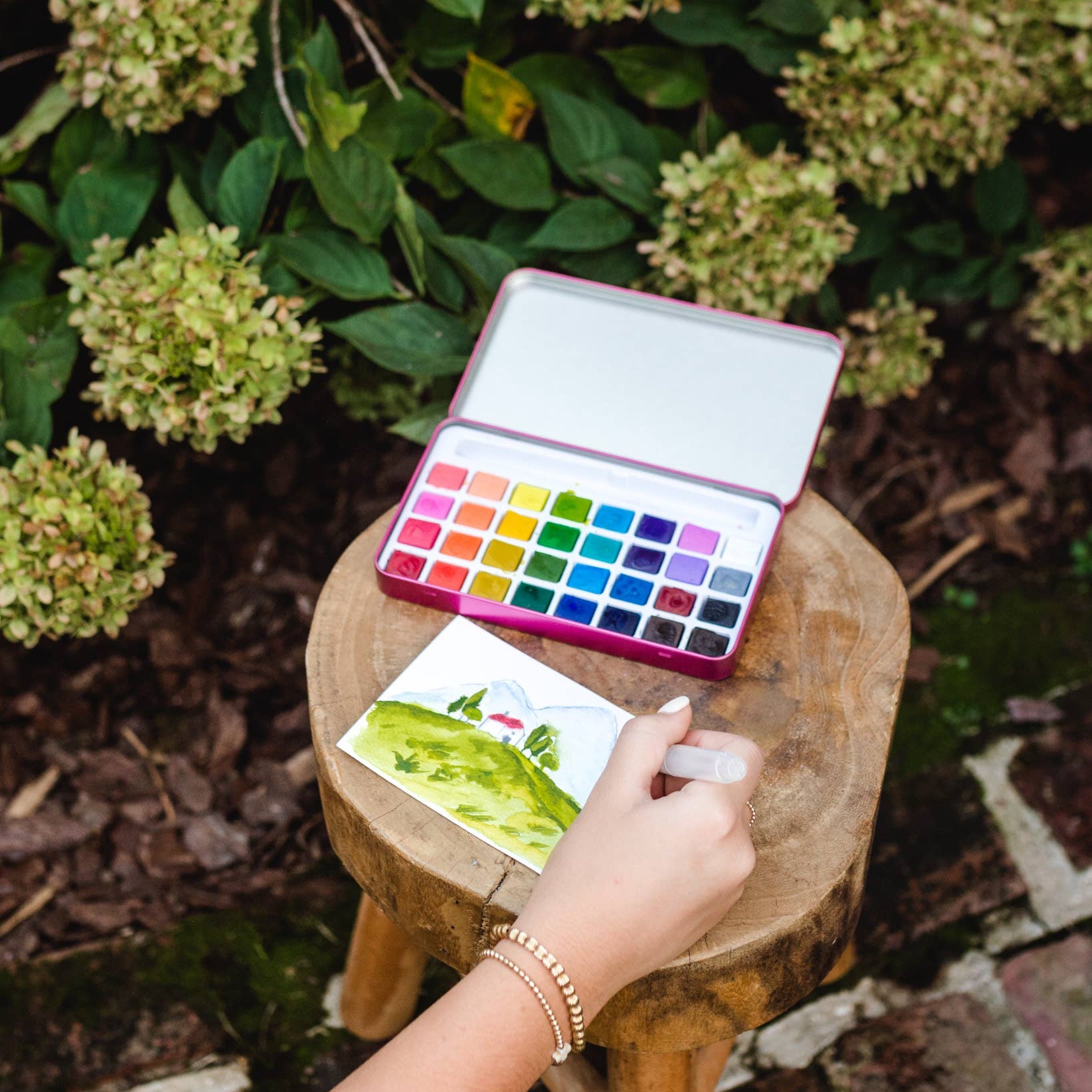 Colorful watercolor palette on a wooden stool with a hand holding a painting, surrounded by greenery.