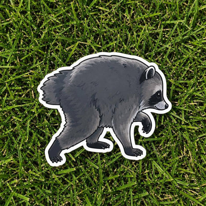 A die-cut vinyl sticker of a walking raccoon, shown lying on a green grass background.