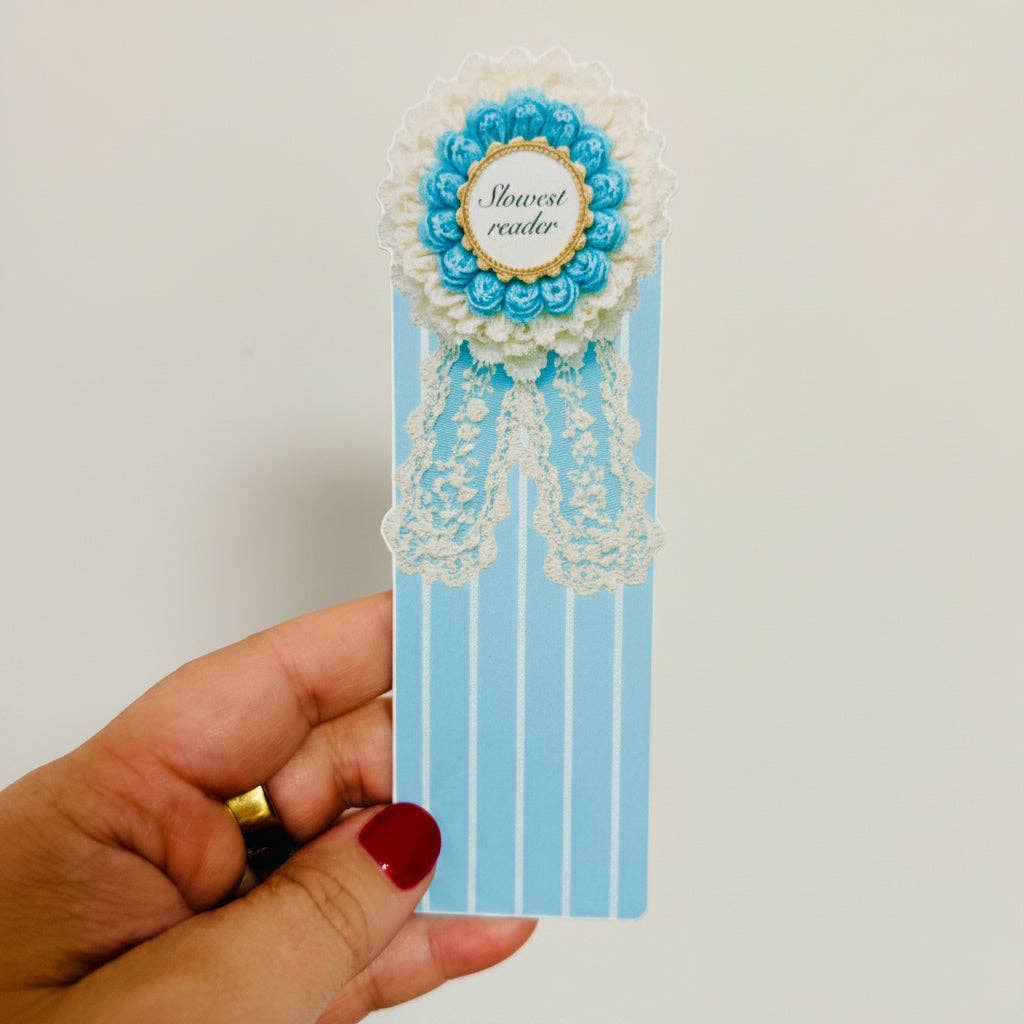 A hand holding a decorative book bookmark featuring light blue and white vertical stripes, topped with a rosette-style floral emblem in blue, white, and gold, and a lace ribbon detail. The center of the emblem reads 'flowerist reader'.
