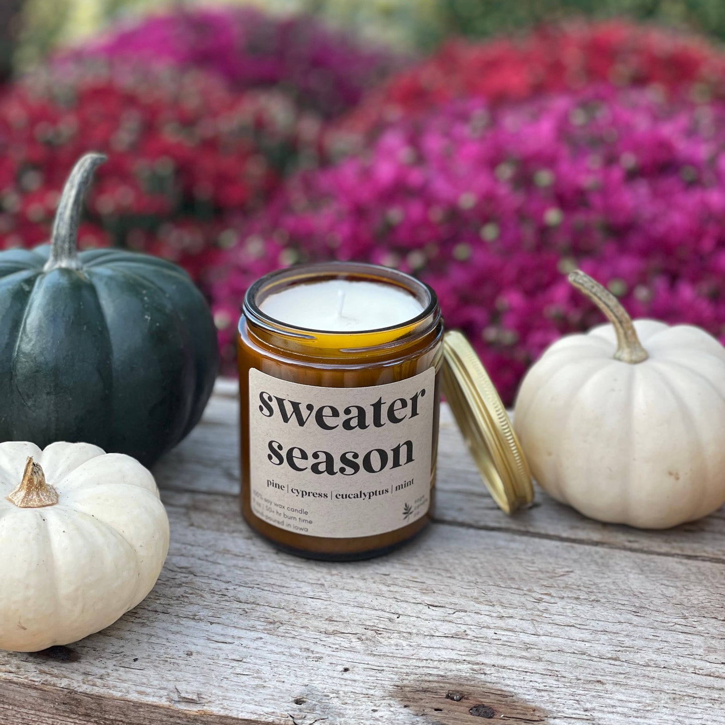 A candle in an amber glass jar with a gold lid, labeled 'sweater season', styled on a rustic wooden surface with small pumpkins and a blurred background of colorful fall flowers.