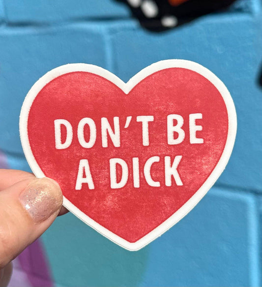 Heart-shaped sticker with 'DON'T BE A DICK' text held by a hand against a blue background