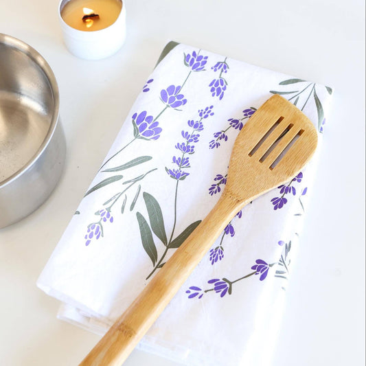 Wooden spatula on a towel with lavender design, next to a metal bowl and candle on a white surface.