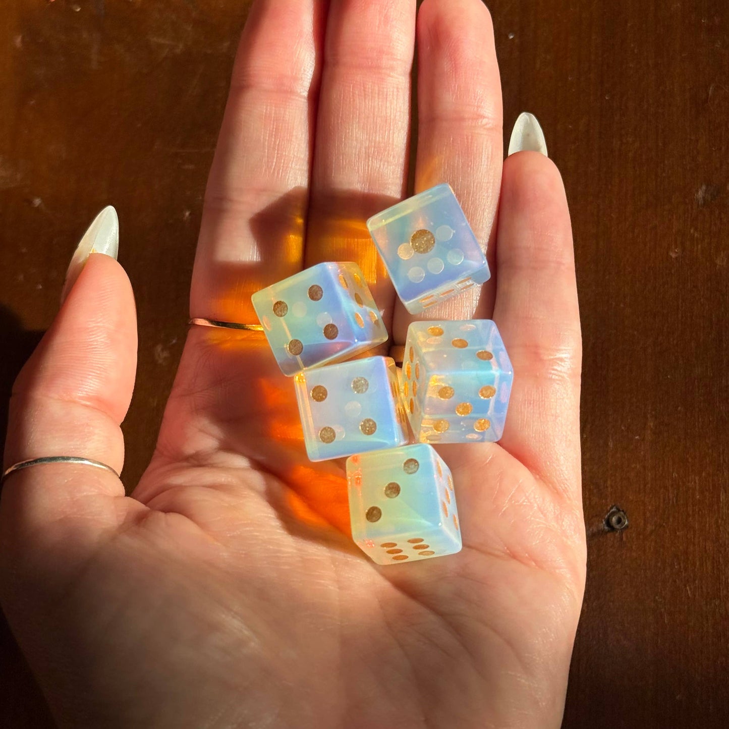 A hand with long manicured nails holds five translucent blue dice with gold dots against a warm brown wooden background.