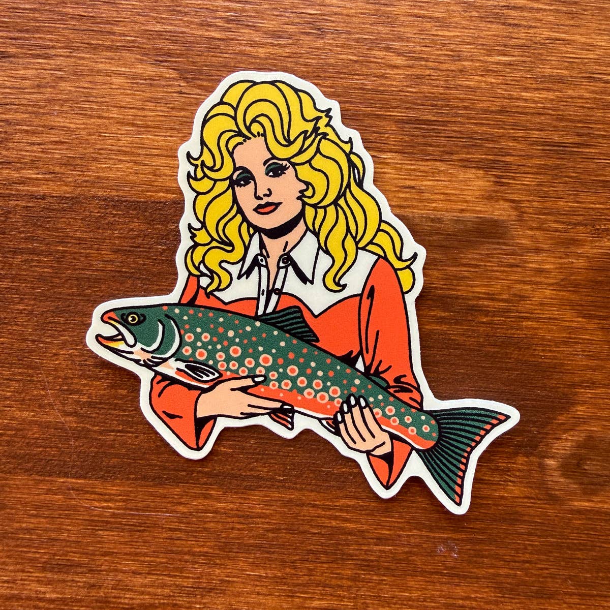 A die-cut vinyl sticker featuring a retro-style illustration of a woman with long blonde hair holding a spotted trout, placed on a brown wooden surface.