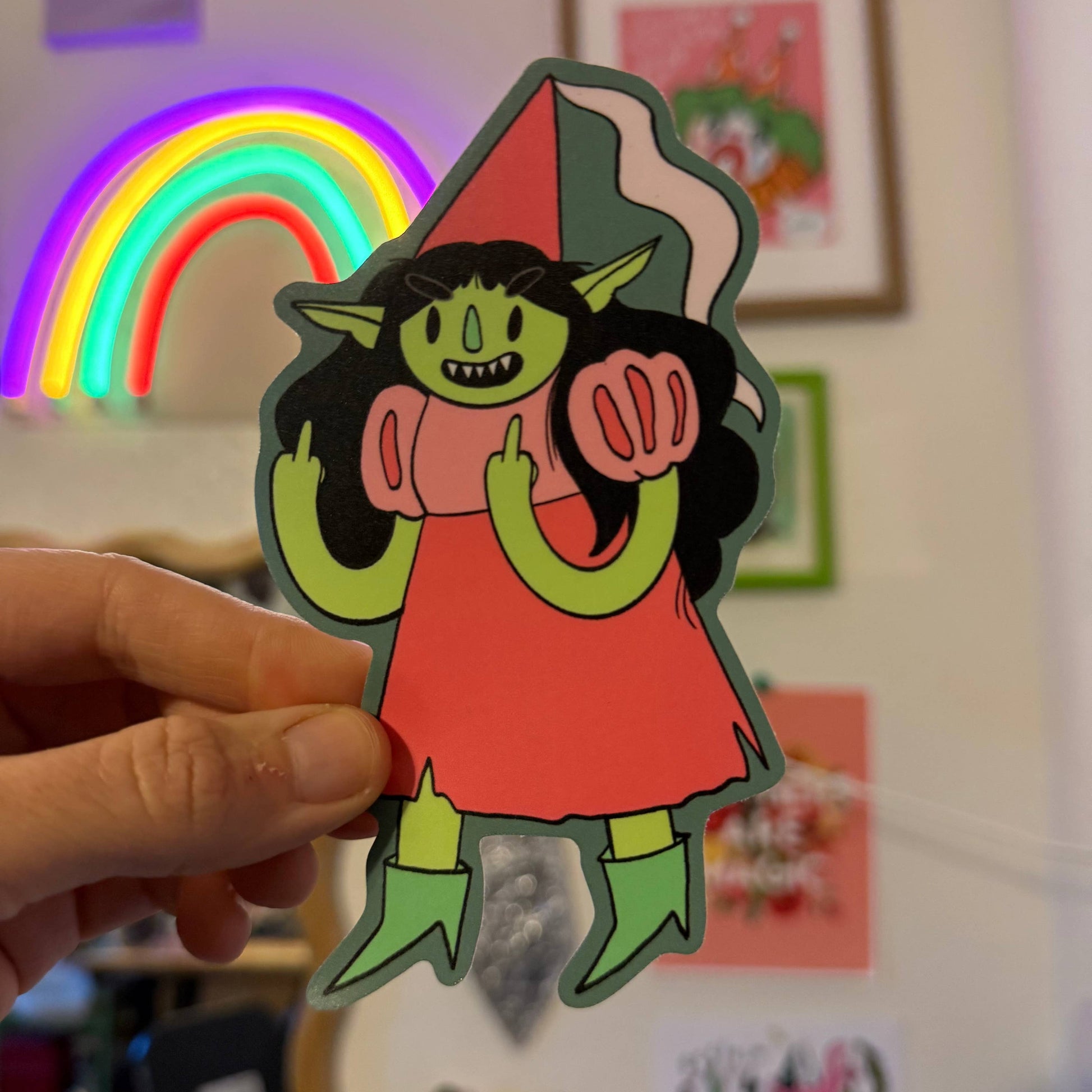 A hand holds a die-cut sticker of a green-skinned elf girl with a red dress and pointed hat against a blurred indoor background with framed art and a neon rainbow.