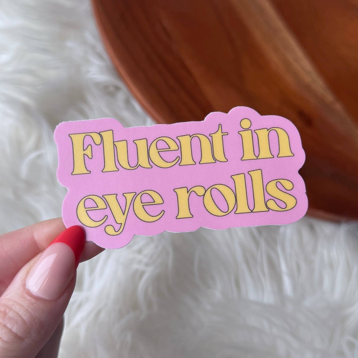 Pink sticker with yellow text 'Fluent in eye rolls' held by a hand against a white background.