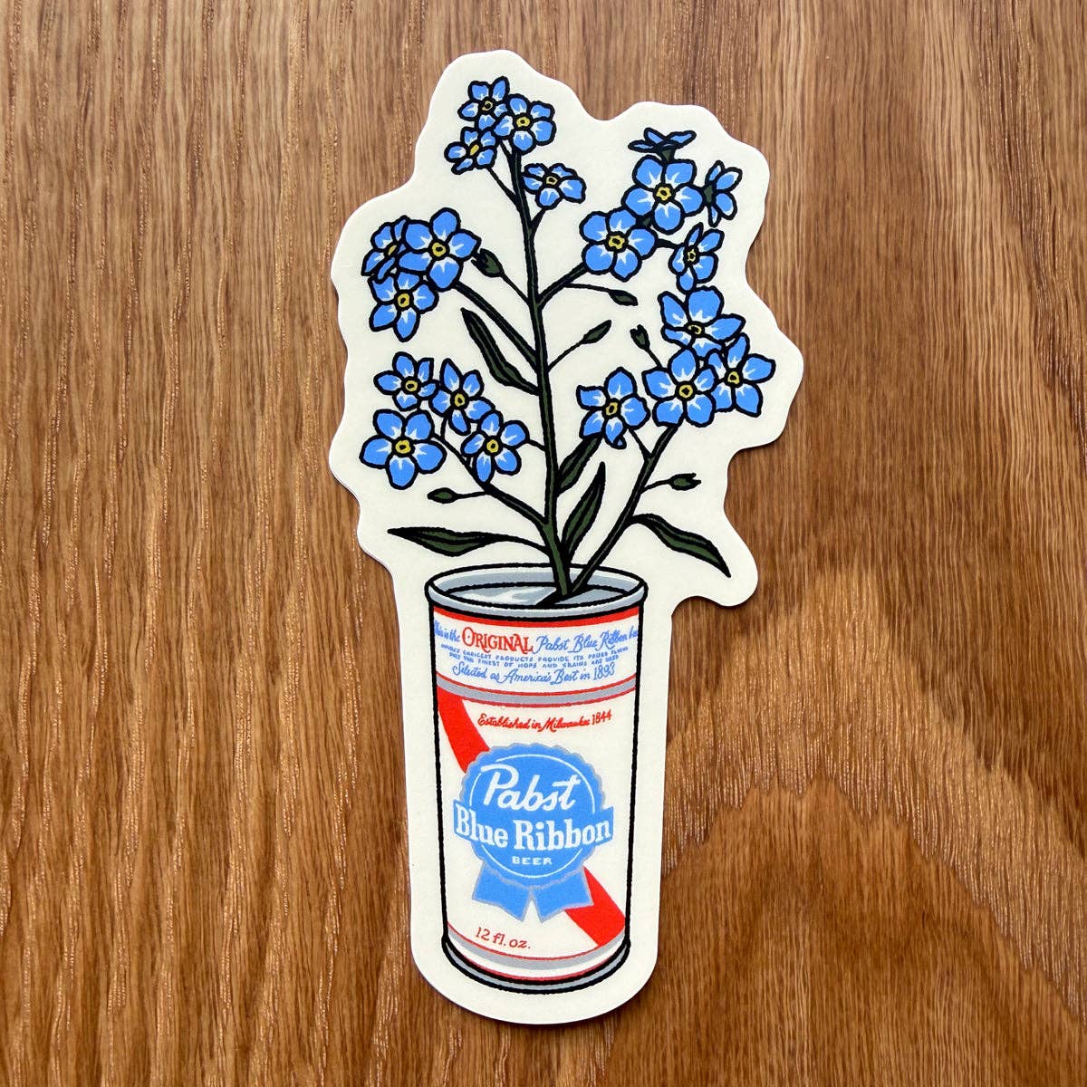 A die-cut sticker featuring blue forget-me-not flowers growing out of a Pabst Blue Ribbon can, placed on a wooden surface.