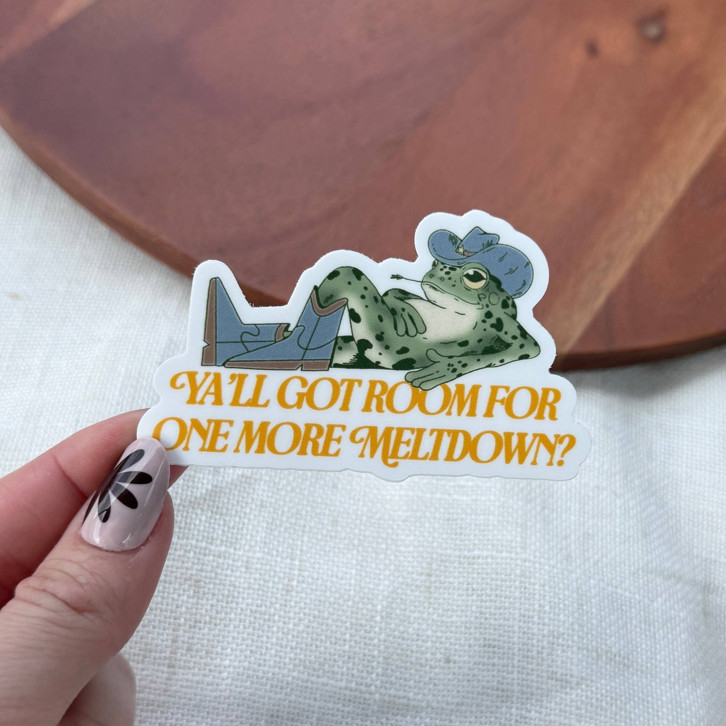 Sticker with a frog design and text held by a hand on a wooden surface.