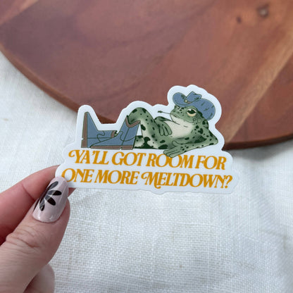 Sticker with a frog design and text held by a hand on a wooden surface.