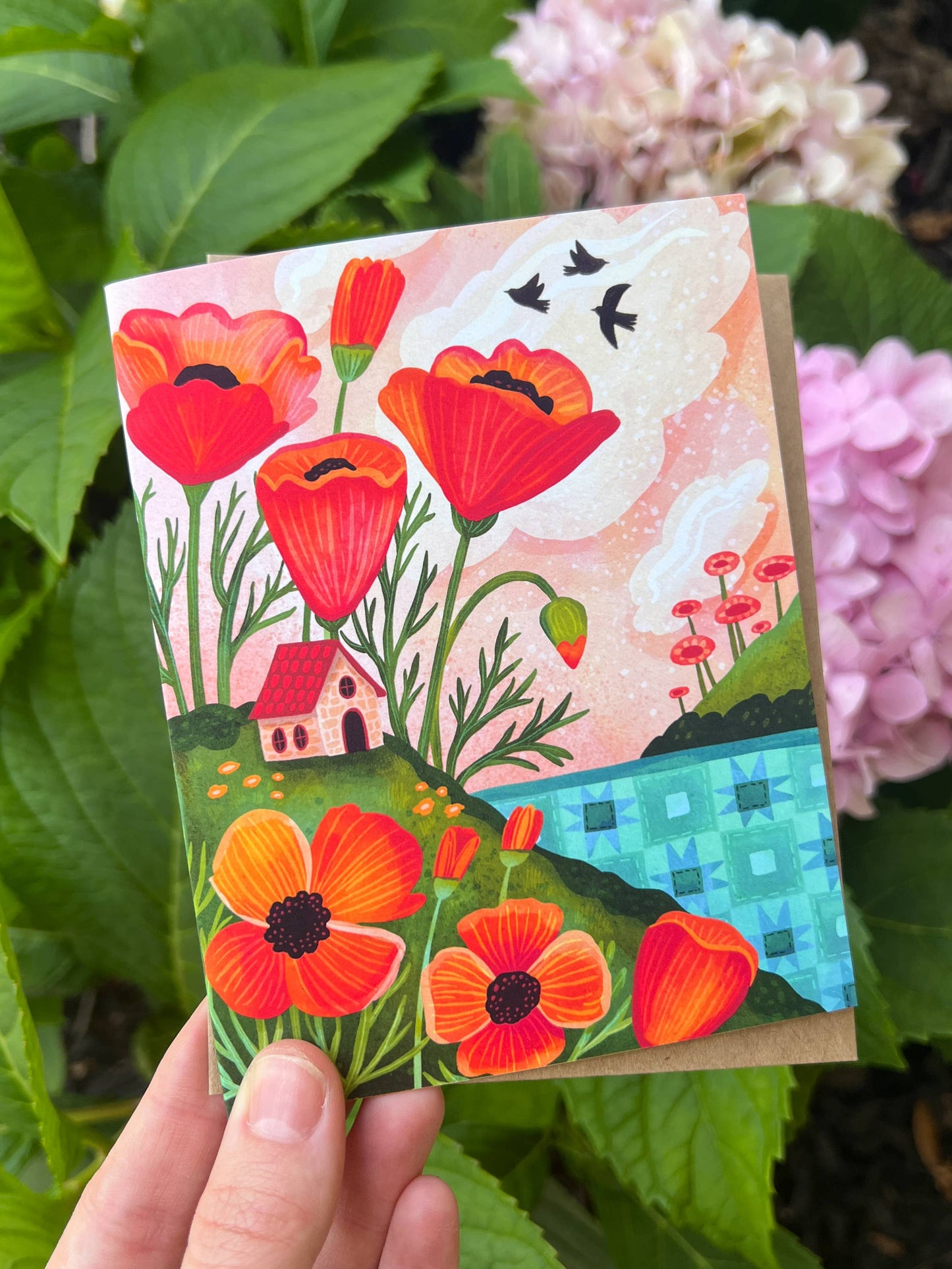 Handheld card with colorful floral design in front of green foliage and pink flowers.