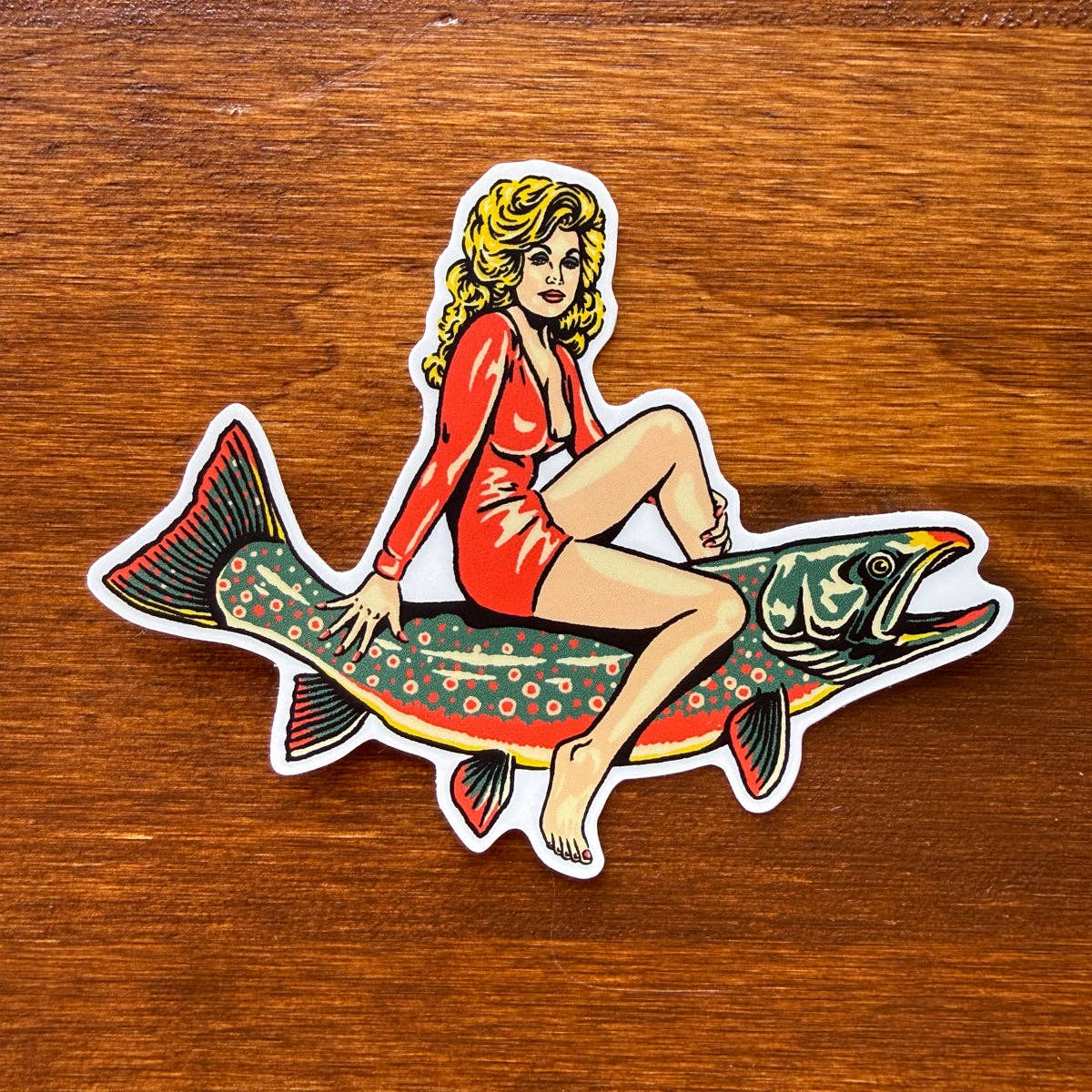 A die-cut sticker featuring a retro-style illustration of a woman in a red swimsuit sitting on a colorful fish, placed on a brown wooden surface.