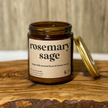 An amber glass jar candle labeled 'rosemary sage' sits on a wooden board with its gold metal lid leaning against it. The background features a light wood wall and a white textured fabric surface.