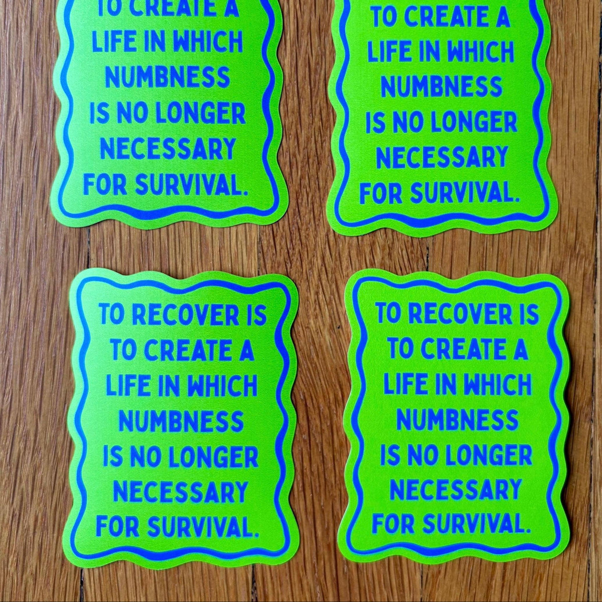 Four identical wavy-edged stickers with the text 'TO RECOVER IS TO CREATE A LIFE IN WHICH NUMBNESS IS NO LONGER NECESSARY FOR SURVIVAL.' printed in blue on a bright green background, arranged on a wooden surface.