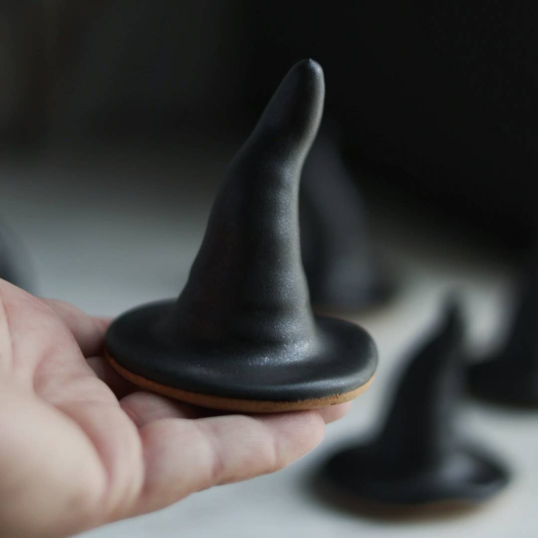 Hand holding a black witch hat cookie with blurred witch hats in the background