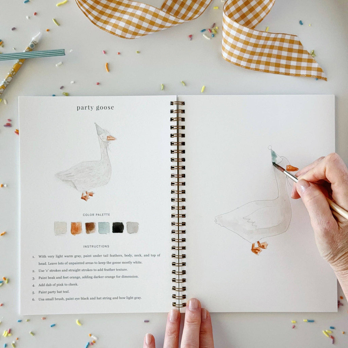 A hand is painting a goose illustration in an open spiral notebook. The left page shows a finished goose drawing, color palette swatches, and step-by-step instructions.