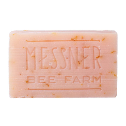 A rectangular bar of Messner Bee Farm soap in a pale peach-pink color, featuring small golden-yellow botanical inclusions and embossed branding on a plain white background.
