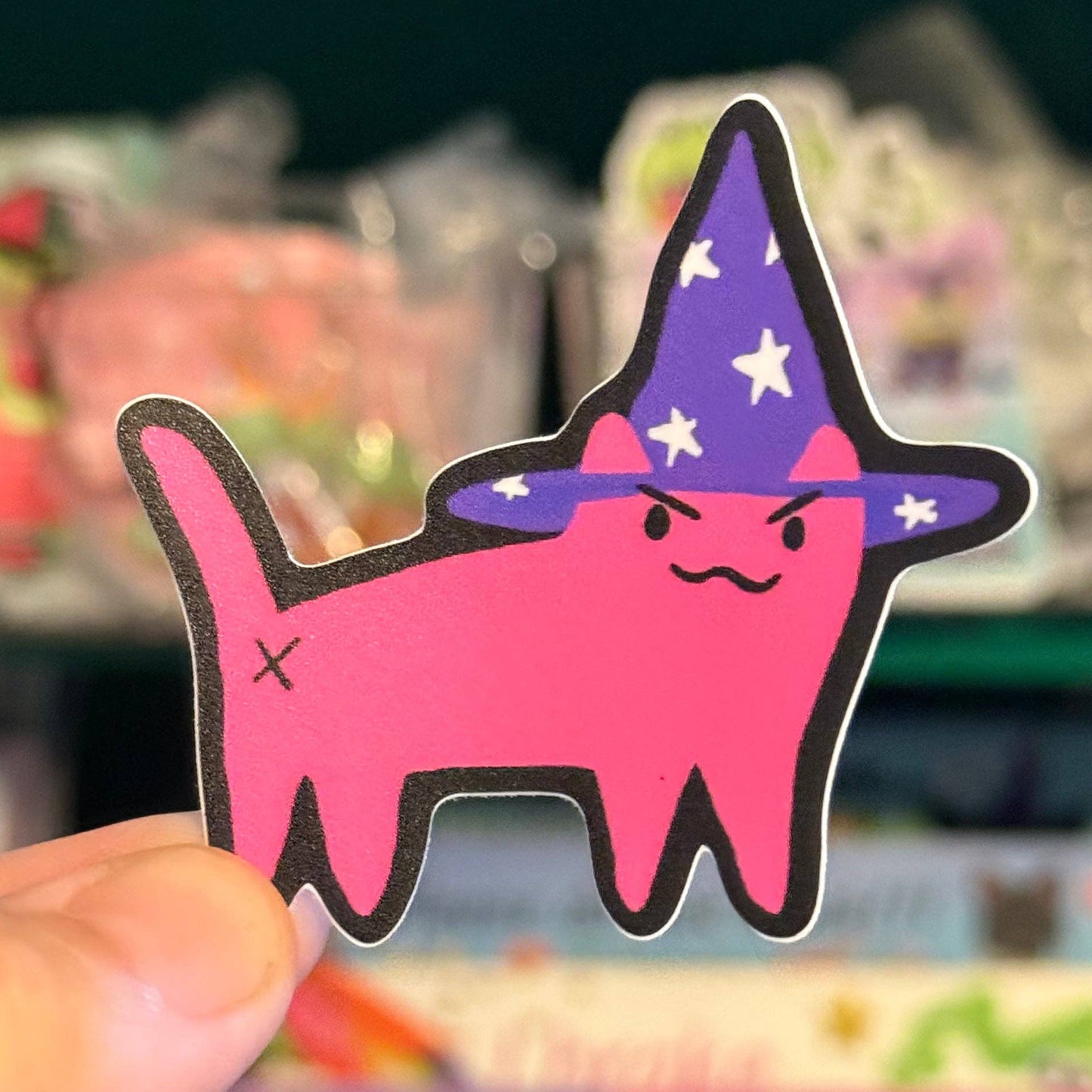 A hand holds a die-cut sticker of a pink cat wearing a purple wizard hat with white stars, set against a blurred store background.