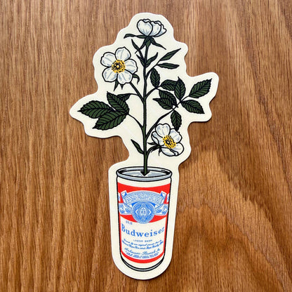 A die-cut sticker featuring white flowers with dark green leaves growing out of a red and blue Budweiser can, placed on a brown wood-grain surface.
