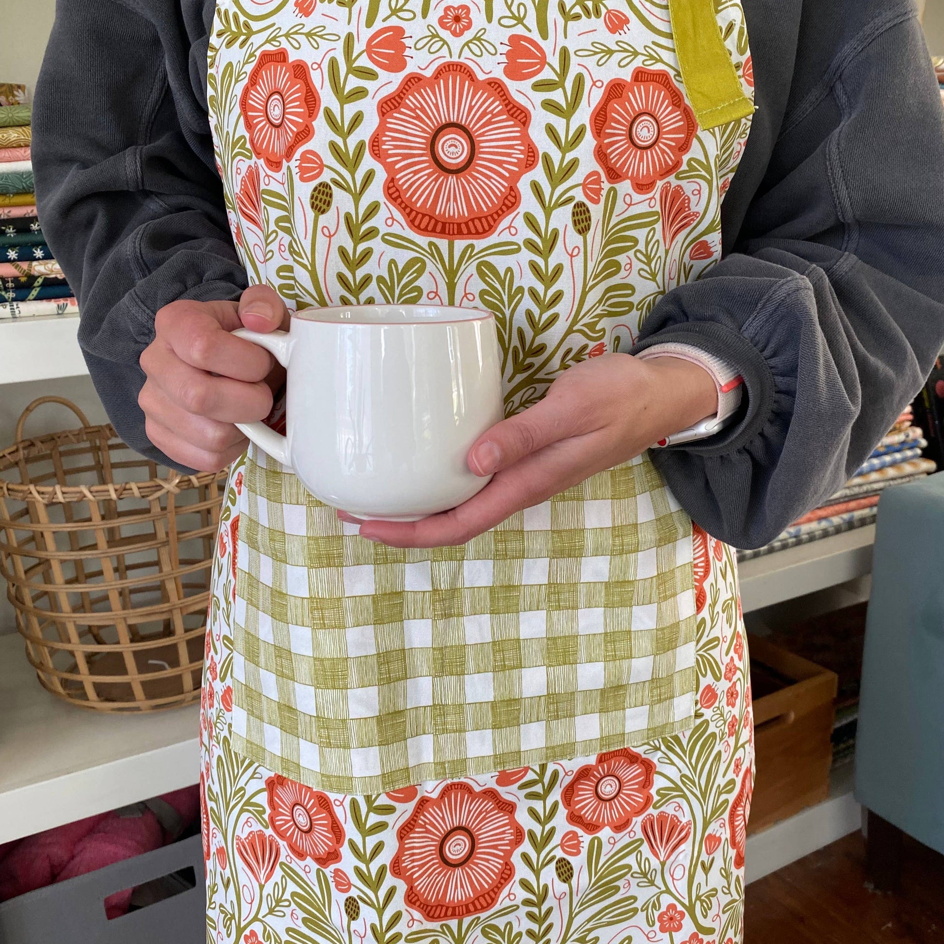 Person wearing a floral apron holding a white mug indoors.