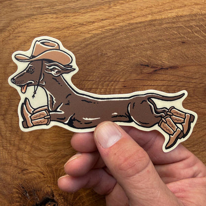 A die-cut sticker of a dachshund wearing a cowboy hat and boots, held by a person's hand over a wooden surface.