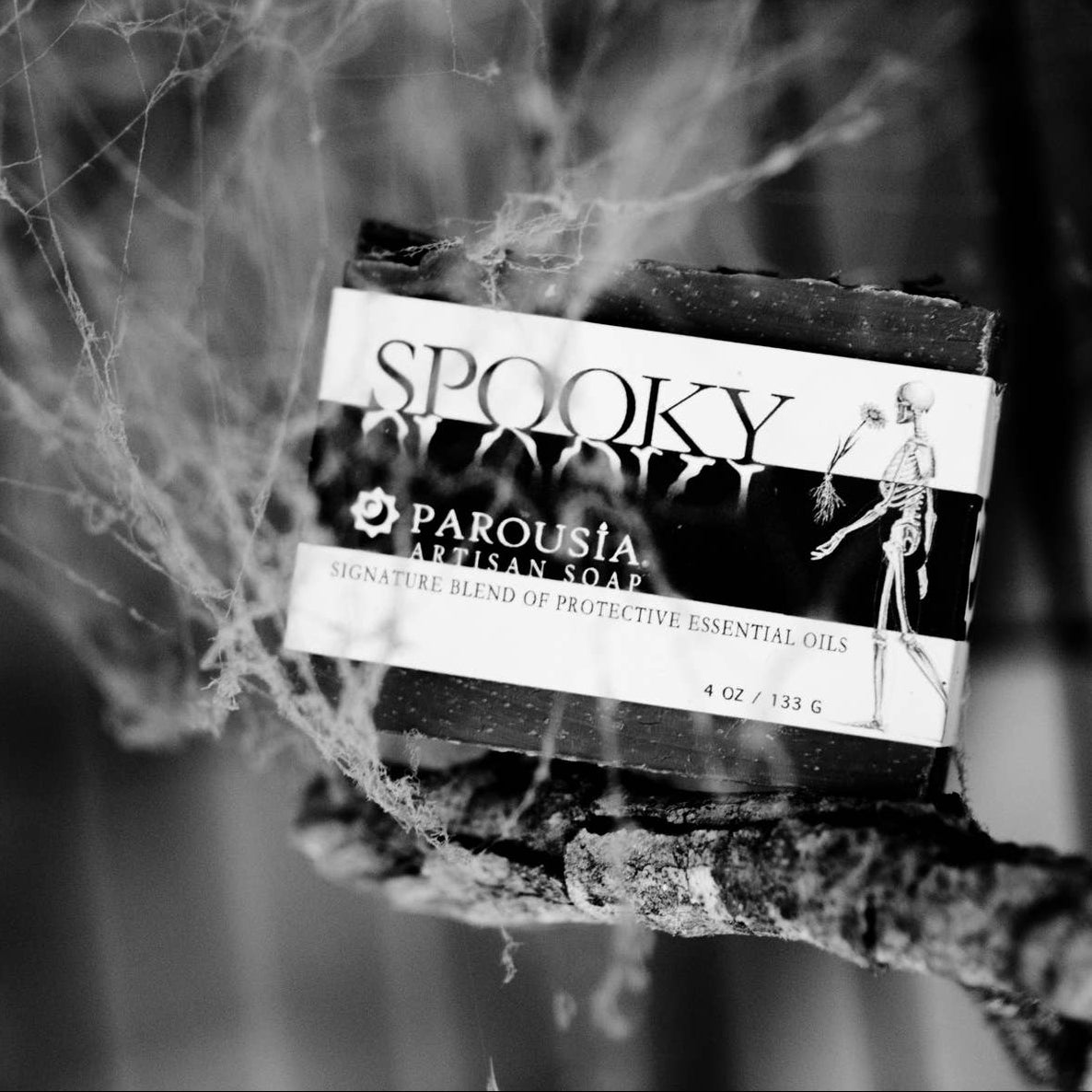 A boxed essential oil product labeled 'SPOOKY' and 'PAROUSIA' resting on a branch, surrounded by spiderwebs in a dark, moody setting.