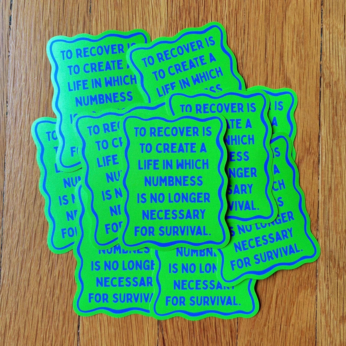 A stack of die-cut stickers with a wavy border, featuring the text 'TO RECOVER IS TO CREATE A LIFE IN WHICH NUMBNESS IS NO LONGER NECESSARY FOR SURVIVAL' in blue lettering on a bright neon green background, displayed on a wooden surface.
