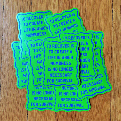 A stack of die-cut stickers with a wavy border, featuring the text 'TO RECOVER IS TO CREATE A LIFE IN WHICH NUMBNESS IS NO LONGER NECESSARY FOR SURVIVAL' in blue lettering on a bright neon green background, displayed on a wooden surface.