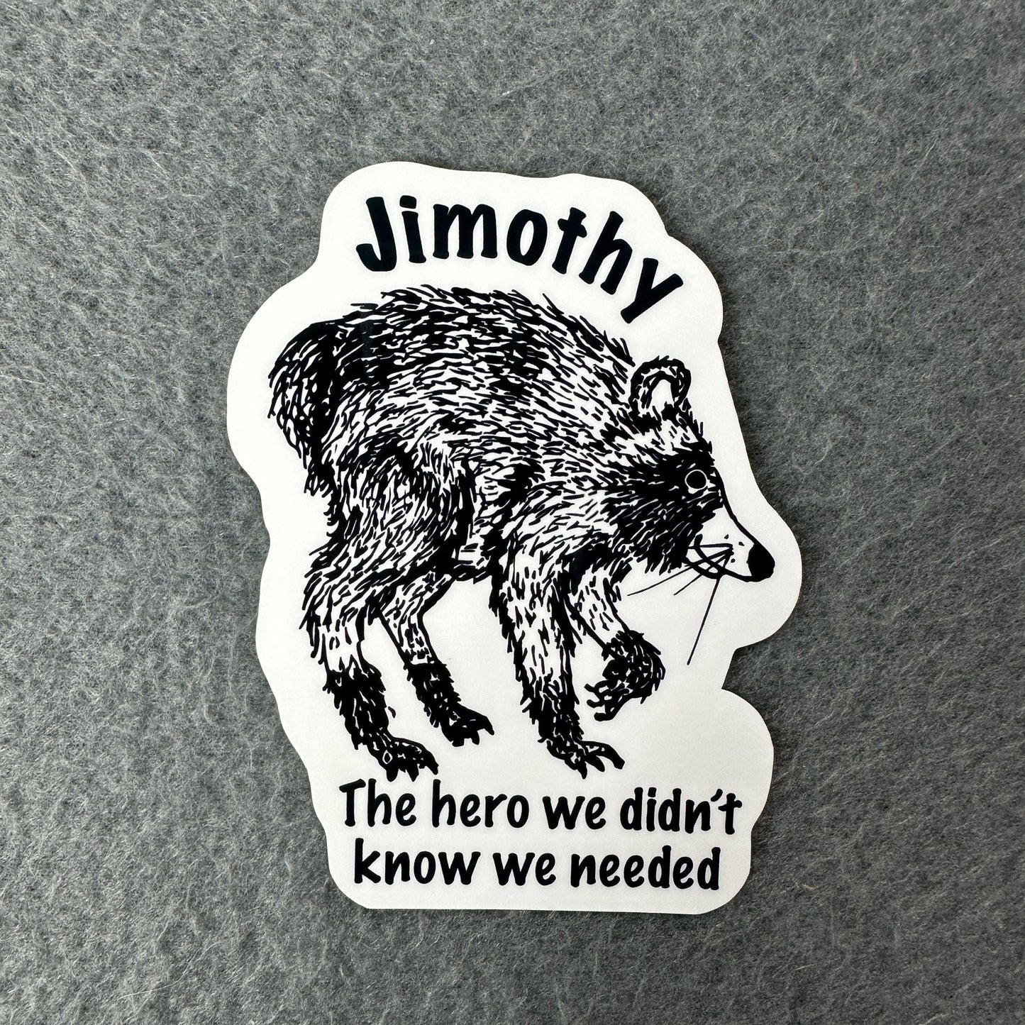A die-cut sticker featuring a black ink sketch of a raccoon on a white background, with the text 'Jimothy' at the top and 'The hero we didn't know we needed' at the bottom, placed on a textured gray surface.