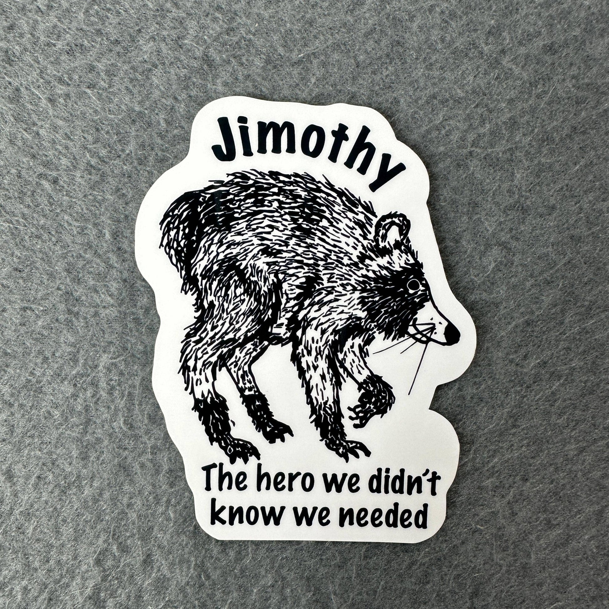 A die-cut sticker featuring a black ink sketch of a raccoon on a white background, with the text 'Jimothy' at the top and 'The hero we didn't know we needed' at the bottom, placed on a textured gray surface.
