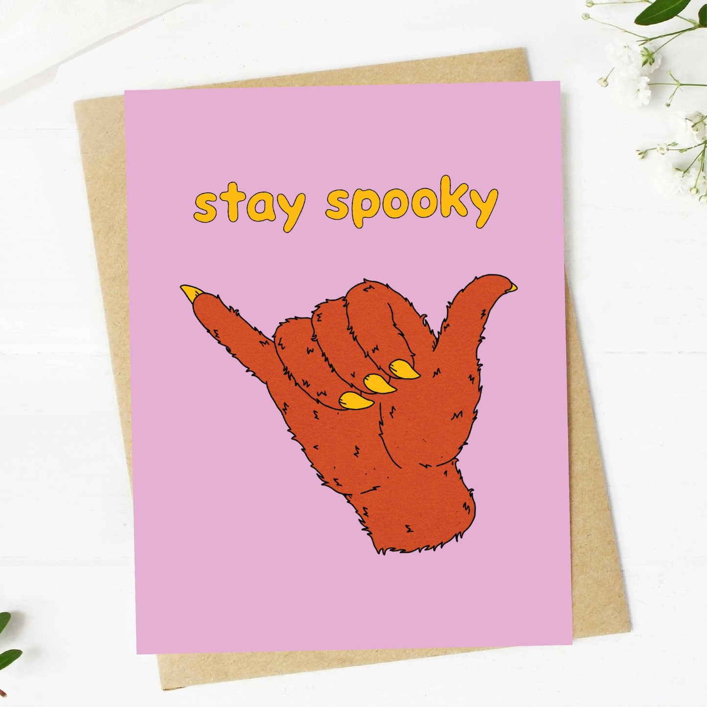 A pink greeting card with the words 'stay spooky' in yellow and an illustrated orange hand making a shaka sign, shown on top of a kraft envelope on a white surface with small floral props.