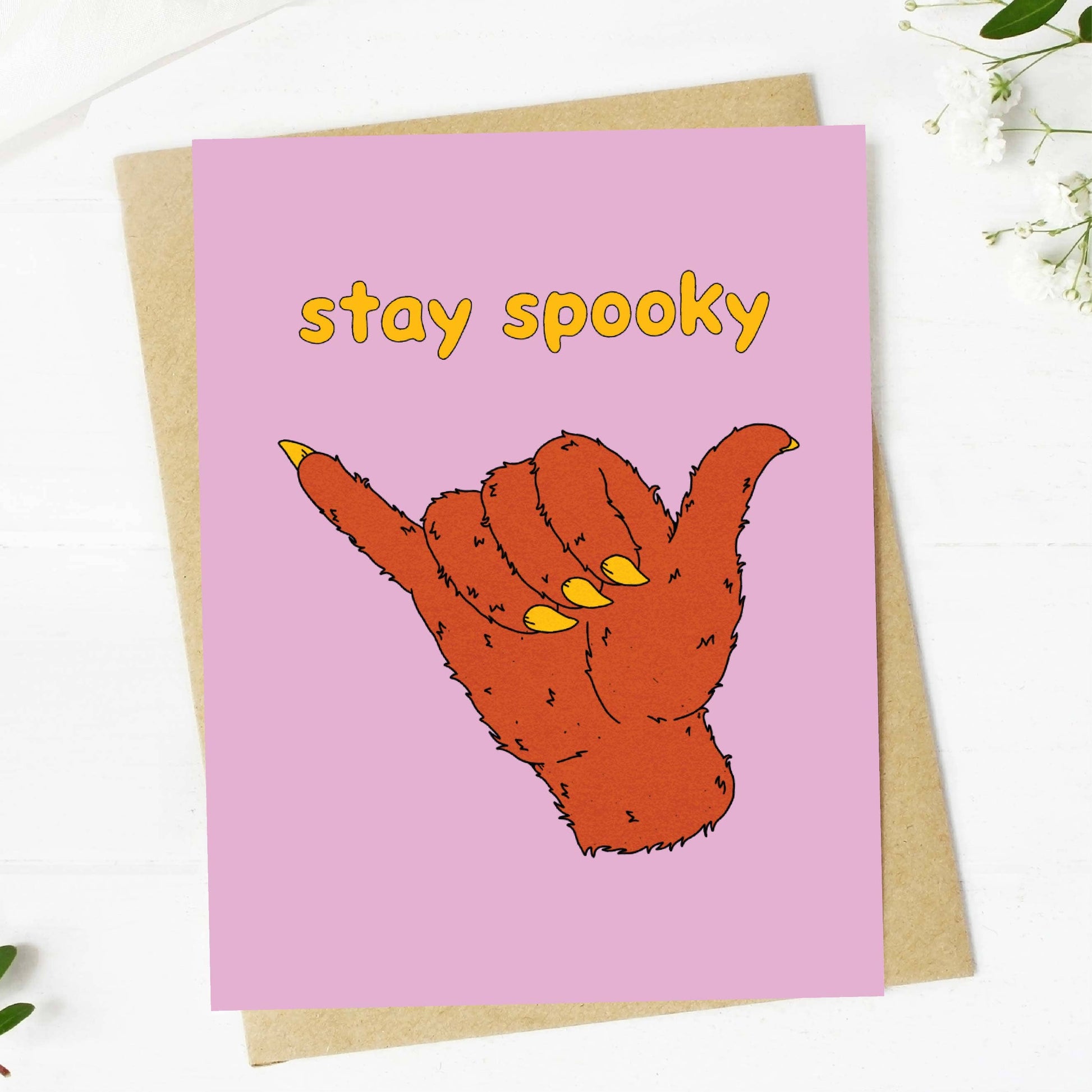 A pink greeting card with the words 'stay spooky' in yellow and an illustrated orange hand making a shaka sign, shown on top of a kraft envelope on a white surface with small floral props.