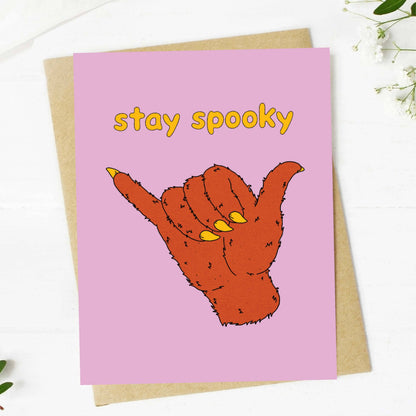 A pink greeting card with the words 'stay spooky' in yellow and an illustrated orange hand making a shaka sign, shown on top of a kraft envelope on a white surface with small floral props.
