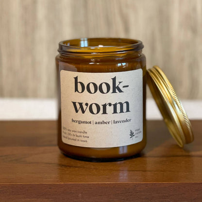 An amber glass jar candle with a gold lid resting beside it on a wooden surface. The front label reads 'book-worm' with smaller scent text below, set against a light wood wall background.