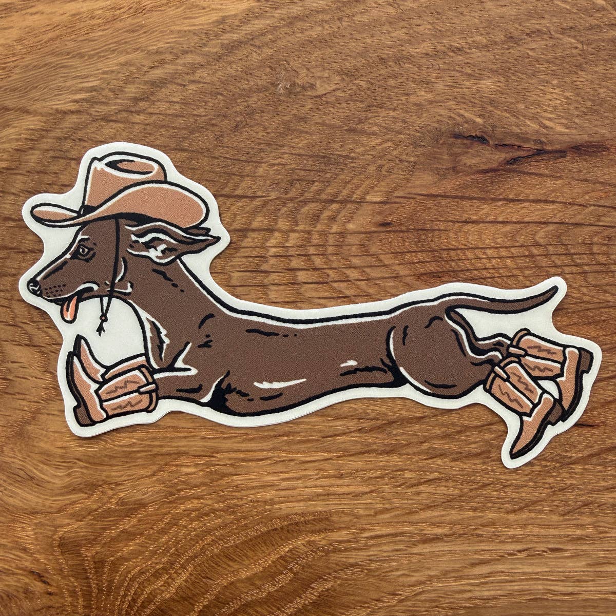 A die-cut vinyl sticker featuring a cartoon illustration of a brown dachshund wearing a tan cowboy hat and boots, placed on a brown wooden surface.
