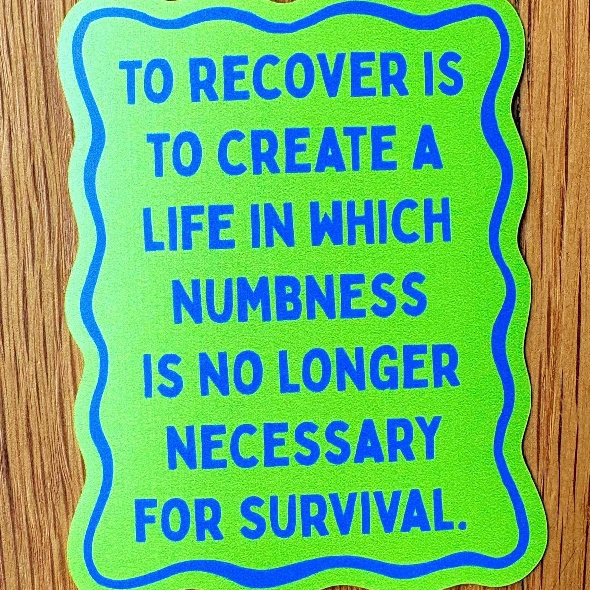 A die-cut sticker with a bright lime green background, blue wavy border, and blue text reading 'TO RECOVER IS TO CREATE A LIFE IN WHICH NUMBNESS IS NO LONGER NECESSARY FOR SURVIVAL.' placed on a brown wood surface.
