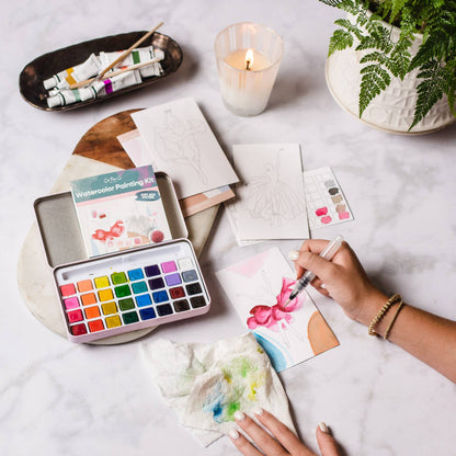 Person painting with watercolors on a table with art supplies and a candle.